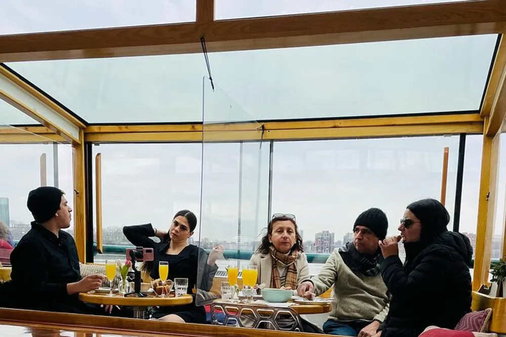 Four individuals are seated at a restaurant table with beverages engaged in conversation in a glass-enclosed space with a city view in the background