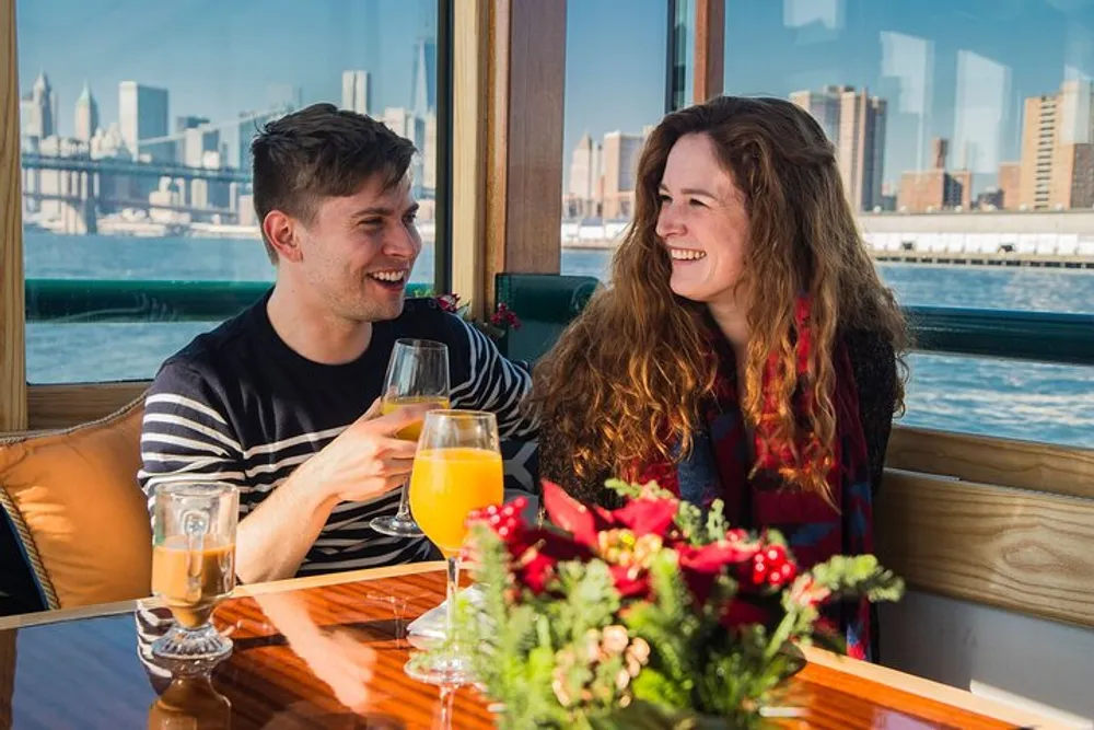 A smiling man and woman are enjoying drinks and a conversation at a table with a city skyline visible through the window behind them