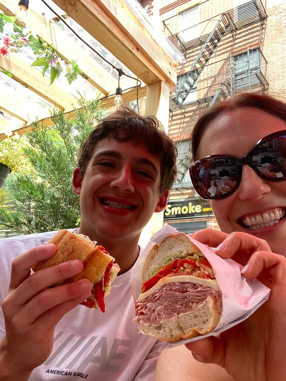 A smiling young man and a woman wearing sunglasses pose with sandwiches in an outdoor setting with a fire escape ladder visible in the background