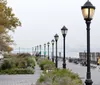 A serene waterfront walkway with lamp posts benches and pedestrians overlooking a river with a bridge in the distant background