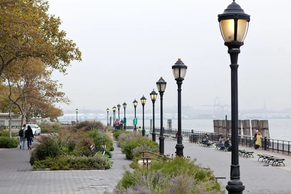 A serene waterfront walkway with lamp posts benches and pedestrians overlooking a river with a bridge in the distant background