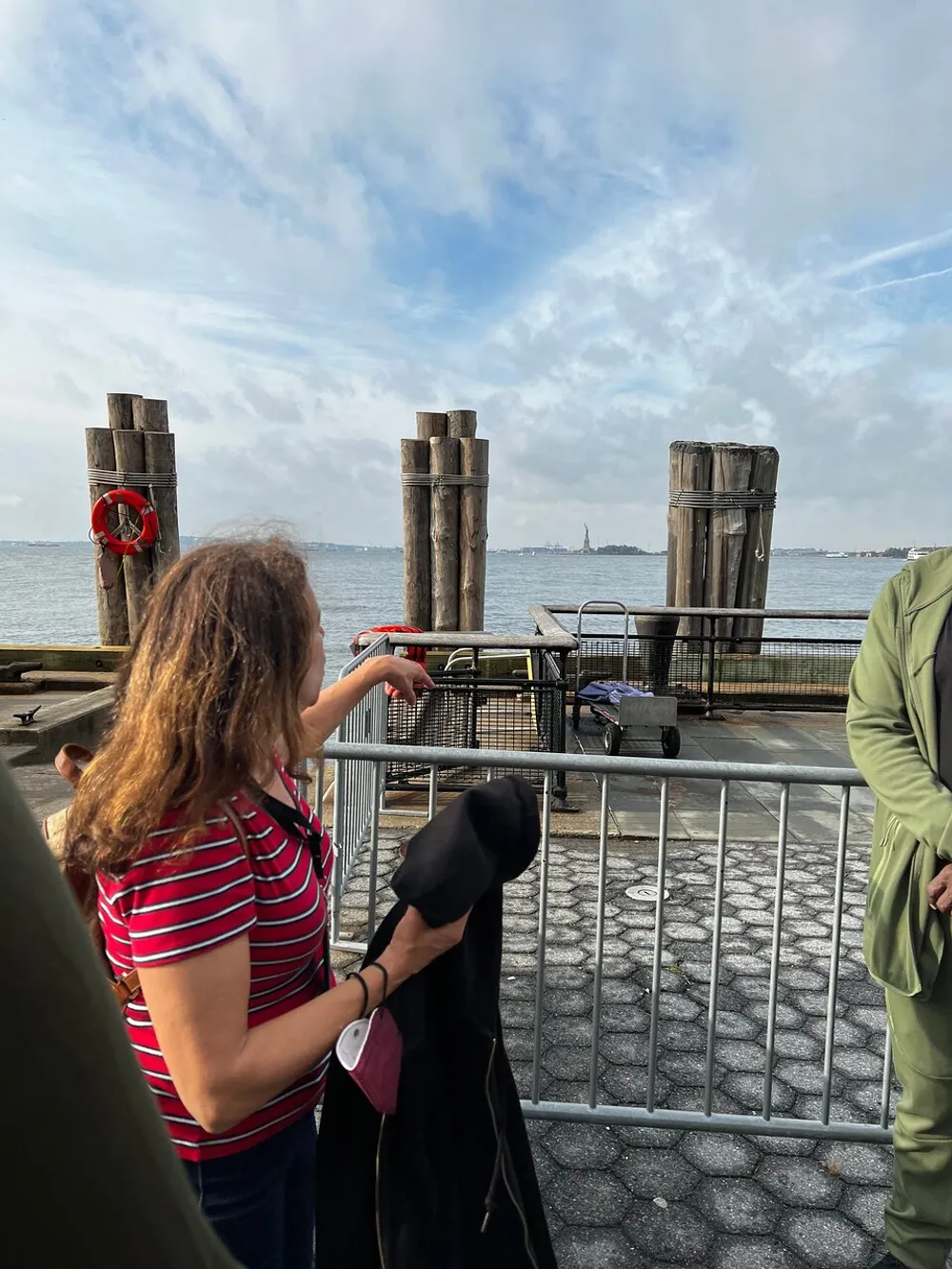 A person is pointing towards the horizon where the Statue of Liberty can be seen in the distance with a waterfront and wooden pilings in the foreground