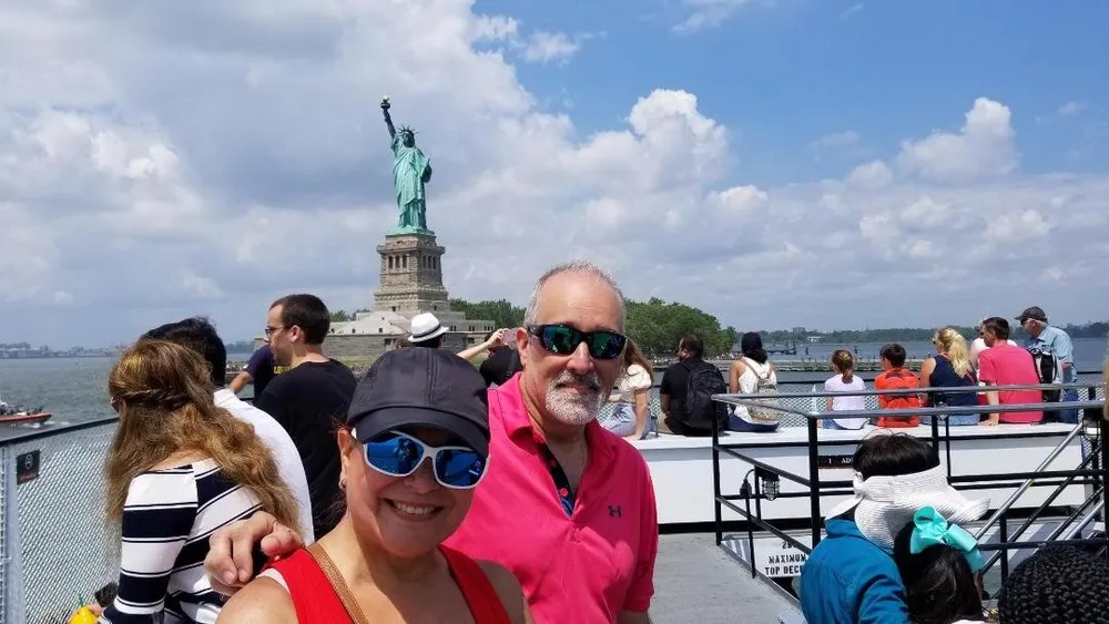 The image features a smiling couple posing for a photo on a boat with the Statue of Liberty in the background surrounded by other passengers enjoying the view