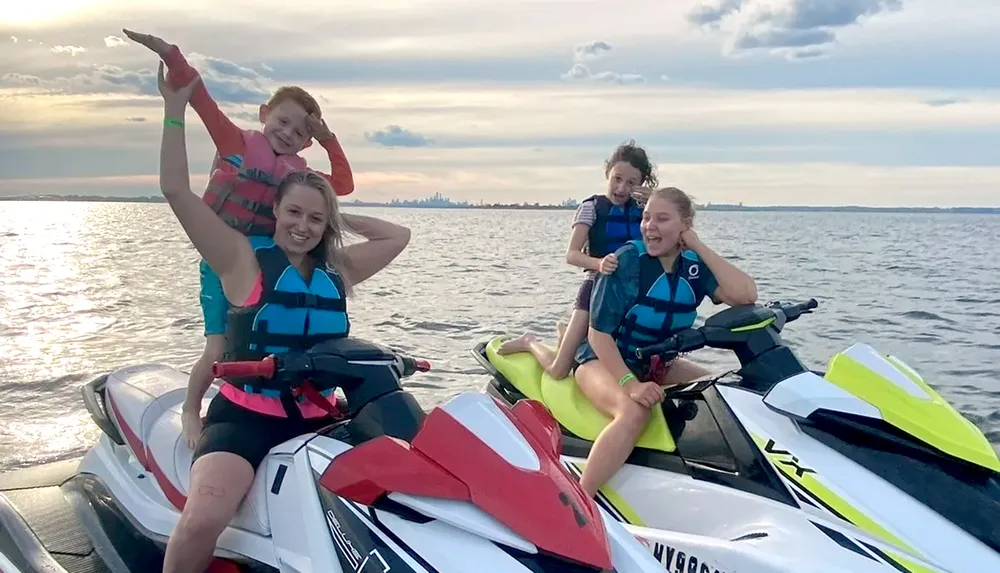 Four people are posing joyfully on two jet skis in calm waters with a cloudy sky above and a distant city skyline on the horizon