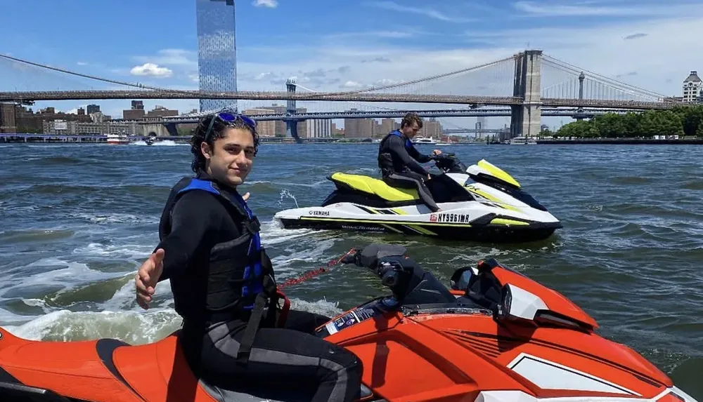 Two people are preparing to ride jet skis on a river with a bridge and skyscrapers in the background
