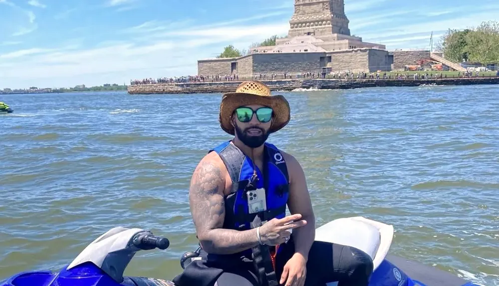 A person wearing a straw hat and sunglasses is posing on a jet ski on the water with a monument in the background