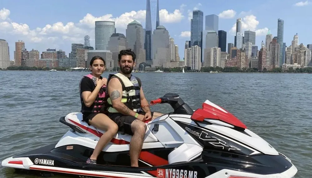 Two people are posing on a jetski with a city skyline in the background