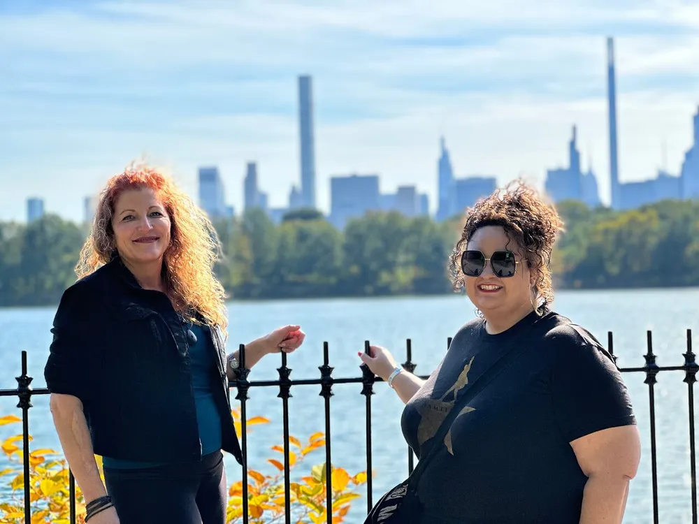 Two smiling people pose in front of a waterscape with a city skyline in the background on a sunny day