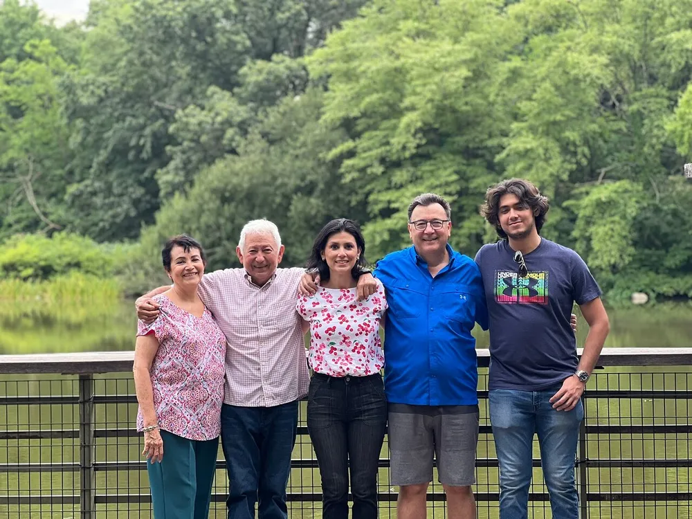 Five people are standing in front of a scenic lush green backdrop smiling and posing together on a bridge with a body of water and vegetation in the background