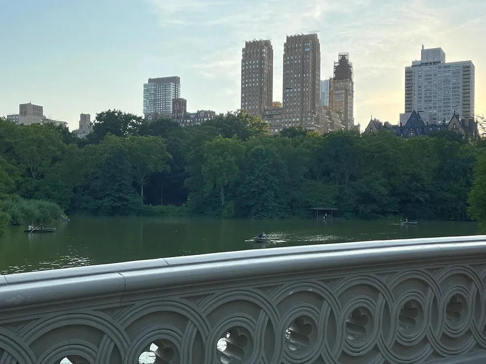 The image depicts a tranquil scene of a lake with people rowing boats framed by an ornate balustrade with a backdrop of dense greenery and urban high-rise buildings