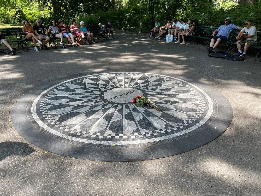 The image shows a mosaic with the word Imagine in the center likely a tribute to John Lennon surrounded by people relaxing on benches in a park-like setting with one person playing a guitar