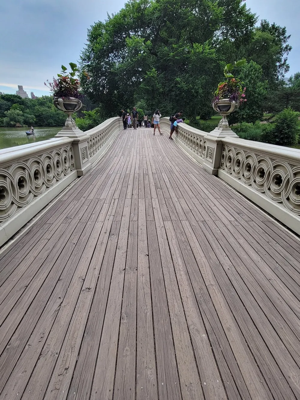 The image depicts a beautifully crafted ornate bridge with a wooden walkway and decorative railings bustling with pedestrians enjoying a leisurely day in a lush park setting