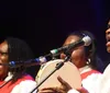 Three people dressed in red and white outfits are singing passionately into microphones with the man on the right also playing a tambourine during what appears to be a spirited live music performance