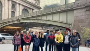 A diverse group of people are posing for a photo in front of the Grand Central Terminal entrance at Pershing Square Plaza.