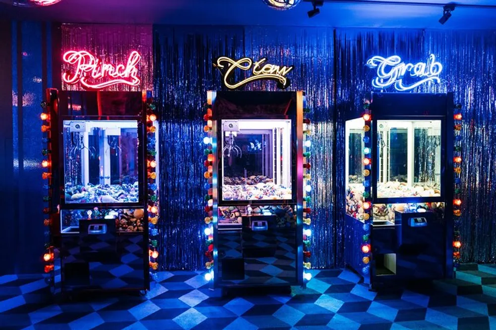 Three claw machines are lit by neon signs in a room with a checkered floor and a metallic fringe backdrop creating a vibrant arcade atmosphere