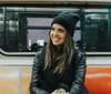 A smiling person wearing a beanie and leather jacket is seated inside a subway car with orange seats holding a phone