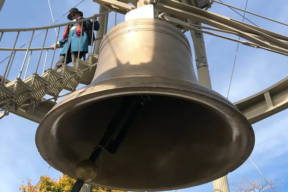 The image shows a large hanging bell with a person standing behind it on an elevated platform set against a clear sky