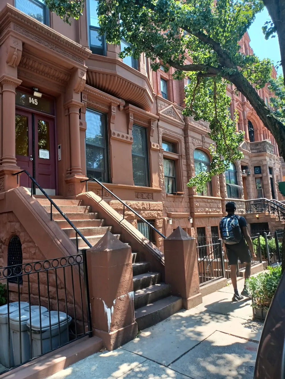A person walks past the entrance to a brownstone building with ornate detailing and a stoop in a tree-lined urban street