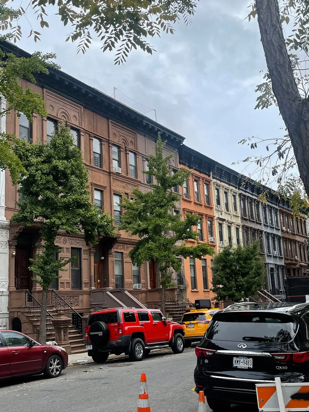 The image shows a street lined with brownstone townhouses a variety of parked cars and some greenery under an overcast sky
