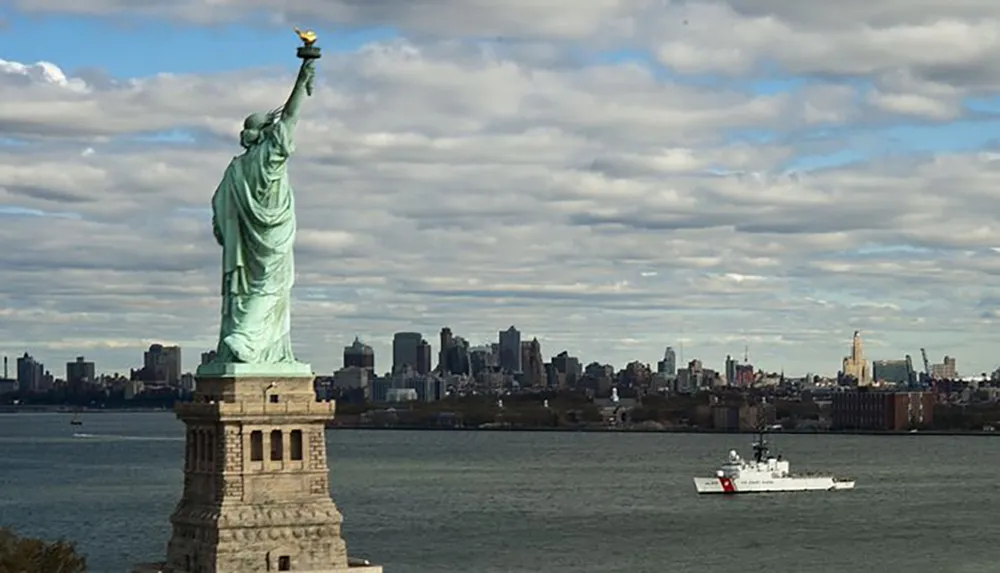The Statue of Liberty stands prominently in the foreground with the New York City skyline and a passing ship in the background