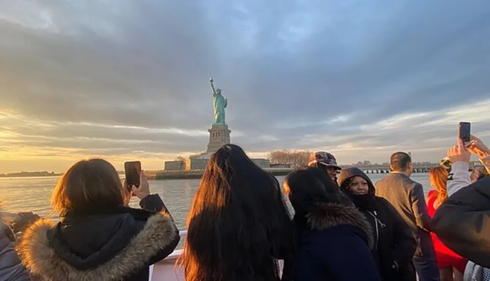 Tourists are capturing the moment on their phones as the sunset illuminates the Statue of Liberty in the background
