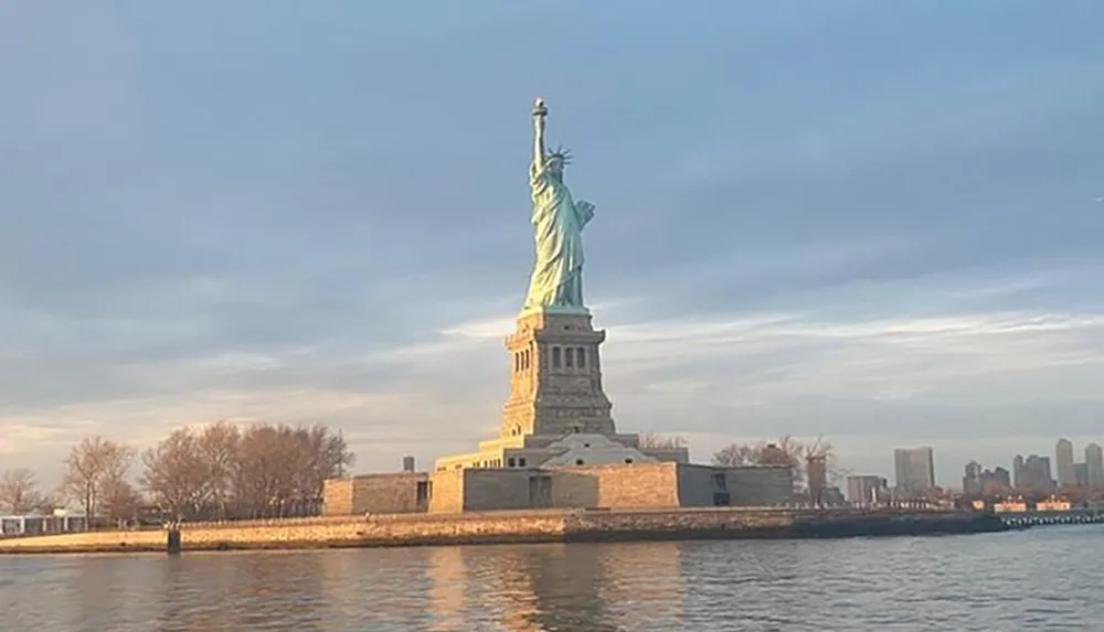 The image shows the Statue of Liberty against a cloudy sky with a soft glow from the sun standing tall on its pedestal near the waters edge