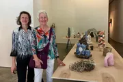 Two women are smiling for a photo while standing next to a table displaying an eclectic array of art pieces in a bright gallery setting.