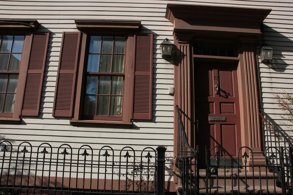 The image shows a section of a buildings facade with two windows with brown shutters siding and a red door accentuated by ornate columns and a wrought iron fence