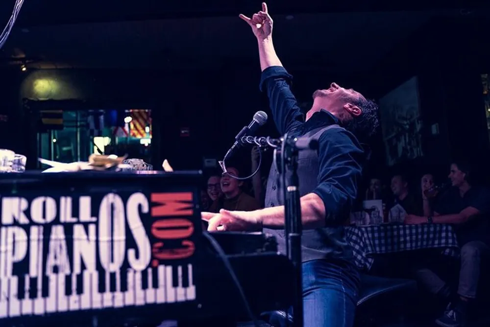 A passionate performer is raising his index finger to the sky while singing into a microphone at a lively piano bar with an audience enjoying the show in the background