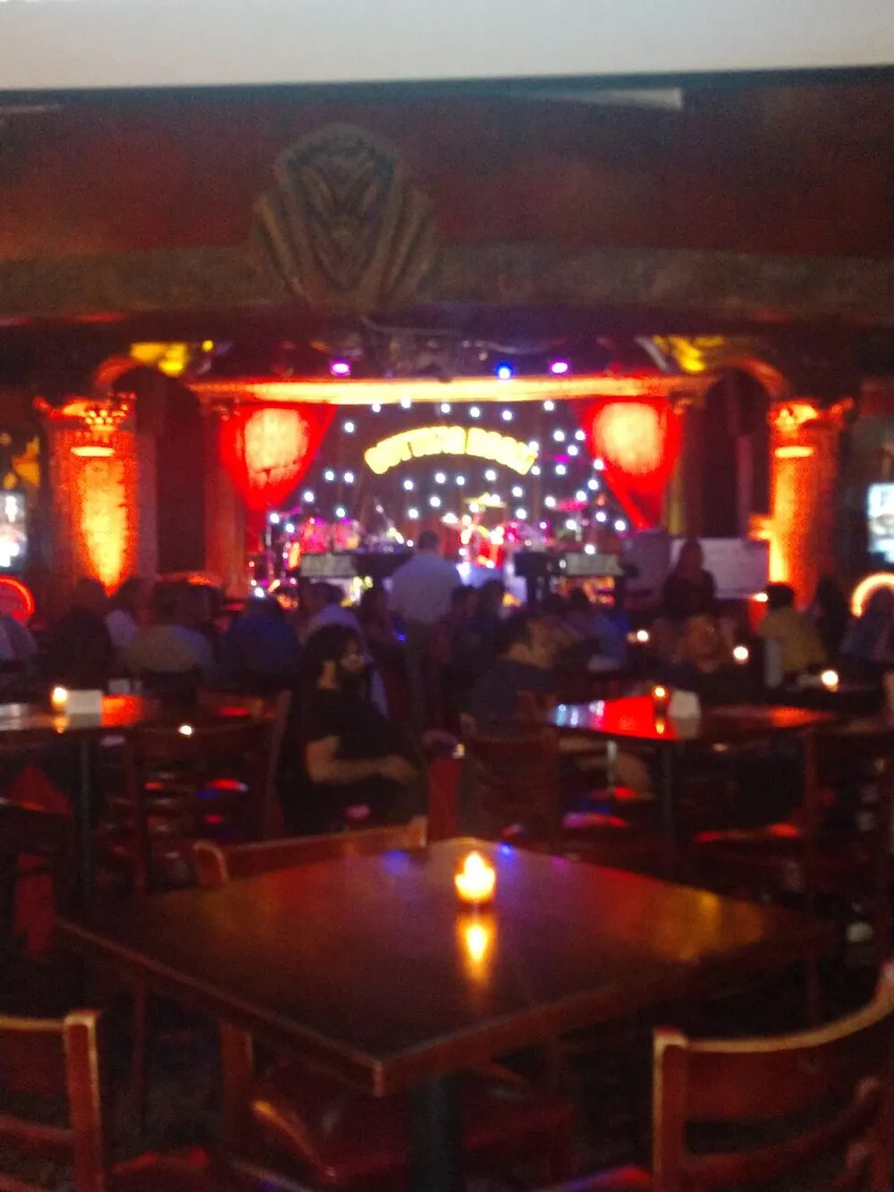 The image shows a dimly lit interior of a venue with people sitting at tables a candlelit atmosphere and a stage with lights and festive decorations in the background