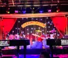 A passionate performer is raising his index finger to the sky while singing into a microphone at a lively piano bar with an audience enjoying the show in the background