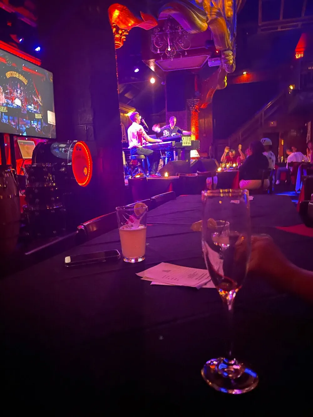 The image depicts a lively atmosphere inside a venue with a dual piano performance a glass of beverage in the foreground and an audience engaged in the entertainment