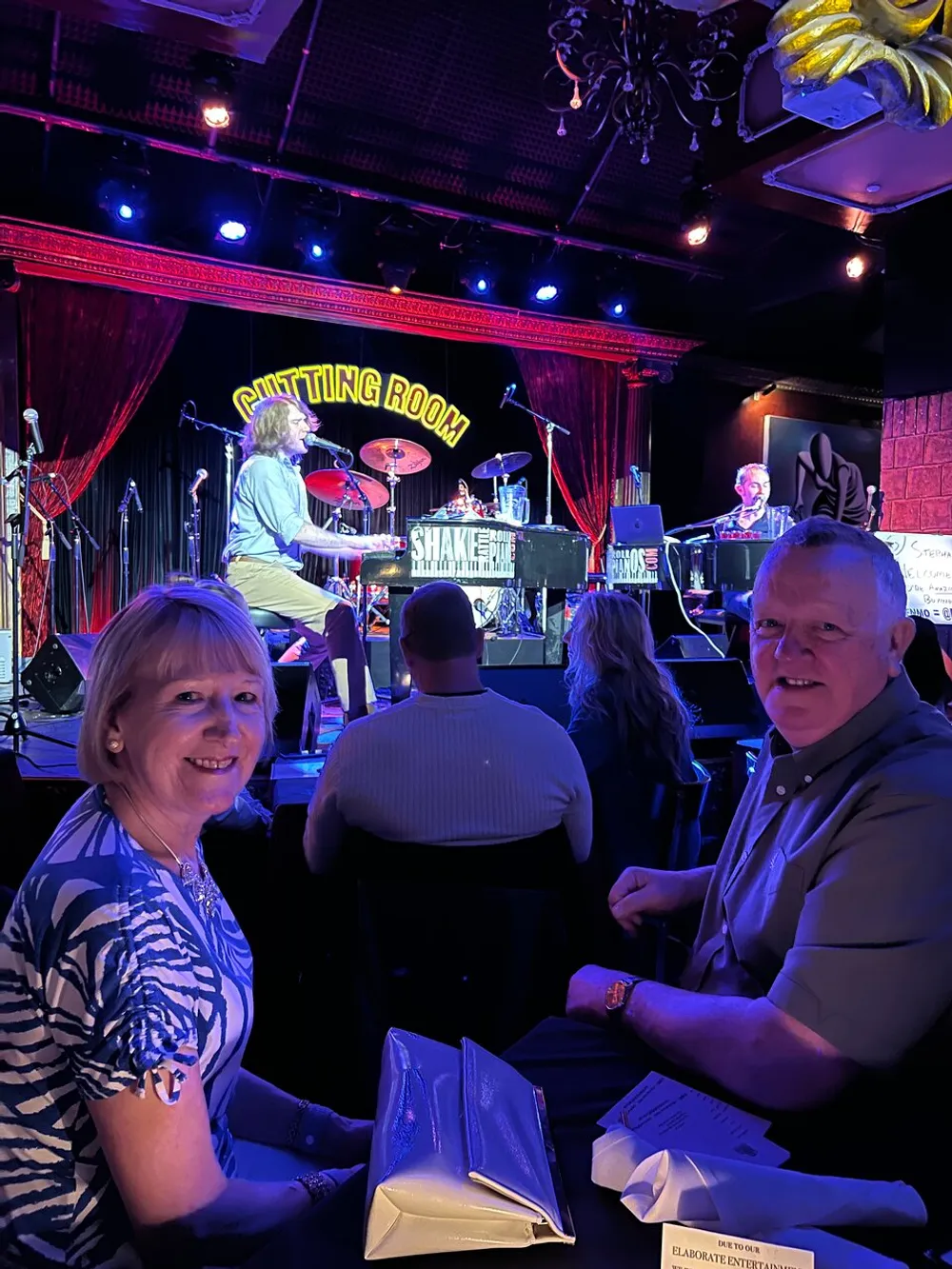 A couple is seated at a table smiling at the camera with a live band performing in the background on a stage with a red curtain and a sign that says The Cutting Room