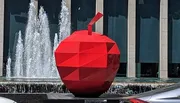 A large red geometric sculpture resembling an apple is placed in front of a fountain with water jets, against a backdrop of a modern building facade.