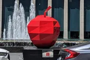 The image shows a large red geometric apple sculpture displayed in an urban setting with a fountain in the background.
