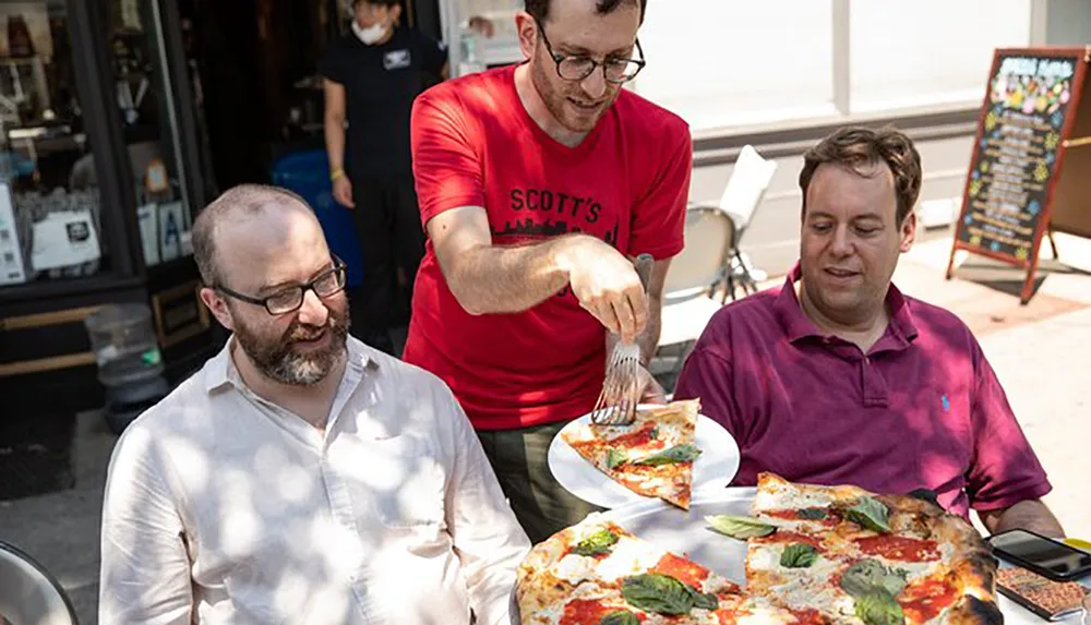 Three people are enjoying a sunny day outdoors with one of them serving a freshly-made large pizza with basil leaves on top