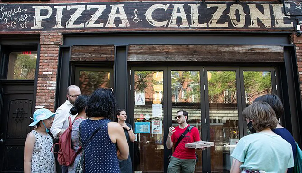 A group of people stands in front of a pizza and calzone restaurant with one person holding a pizza box