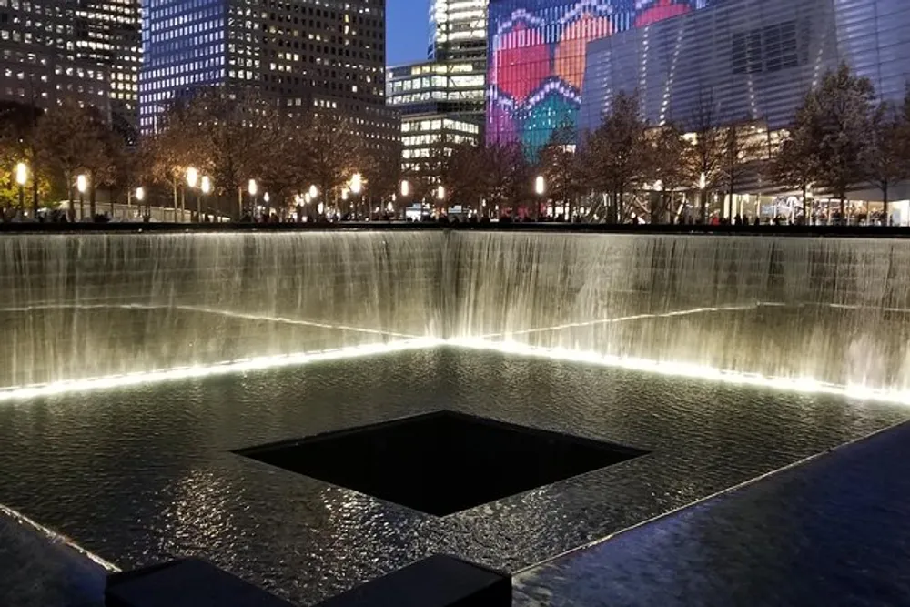 The image depicts a serene evening view of a large reflective memorial pool with cascading waterfalls encircled by trees against a backdrop of illuminated city buildings