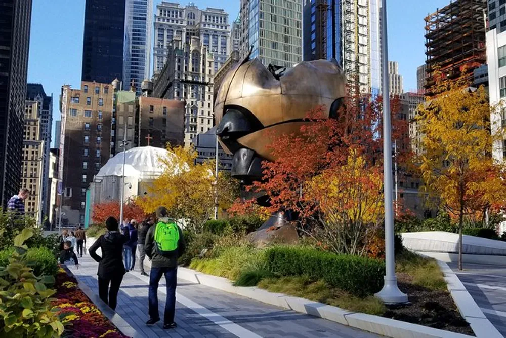 The image shows pedestrians walking through an urban park with colorful autumn foliage with a large abstract bronze sculpture in the background set against a backdrop of towering buildings