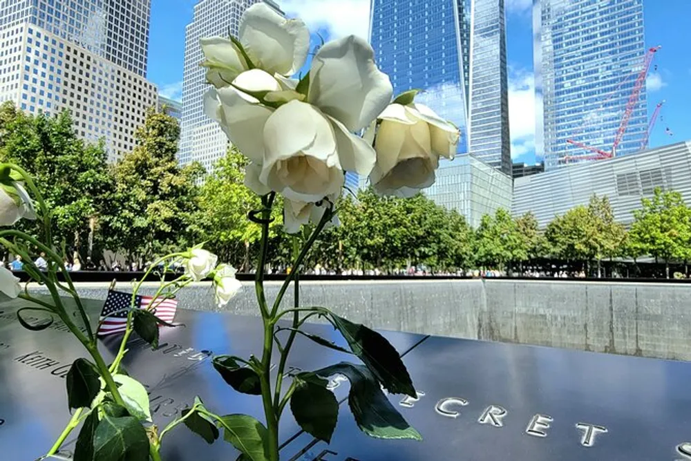 The image shows a close-up view of white roses placed at a memorial site with names inscribed on it with modern buildings and a blue sky in the background