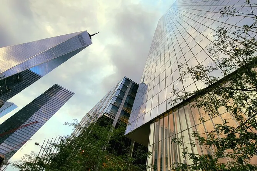 The image captures a dynamic upward view of towering skyscrapers juxtaposed with greenery under a cloudy sky