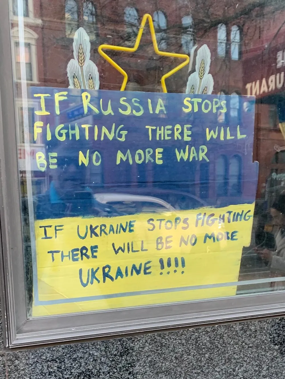 The image depicts a handwritten sign with a statement about the cessation of fighting by Russia leading to no more war and a contrasting statement about Ukraine likely expressing a political viewpoint on an ongoing conflict