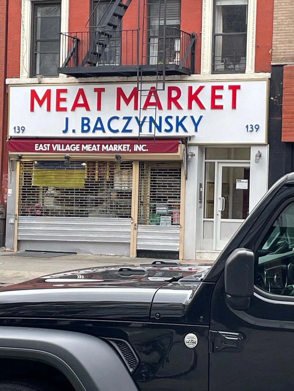 The image shows a storefront with a sign that reads MEAT MARKET J BACZYNSKY above a closed security gate indicating it may be a specialty butchery named after someone with Eastern European heritage located at building number 139