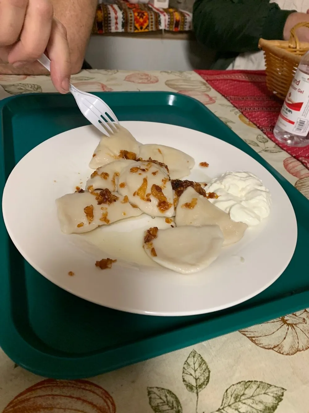A person is about to eat dumplings topped with fried onions and a side of sour cream on a white plate placed on a green tray