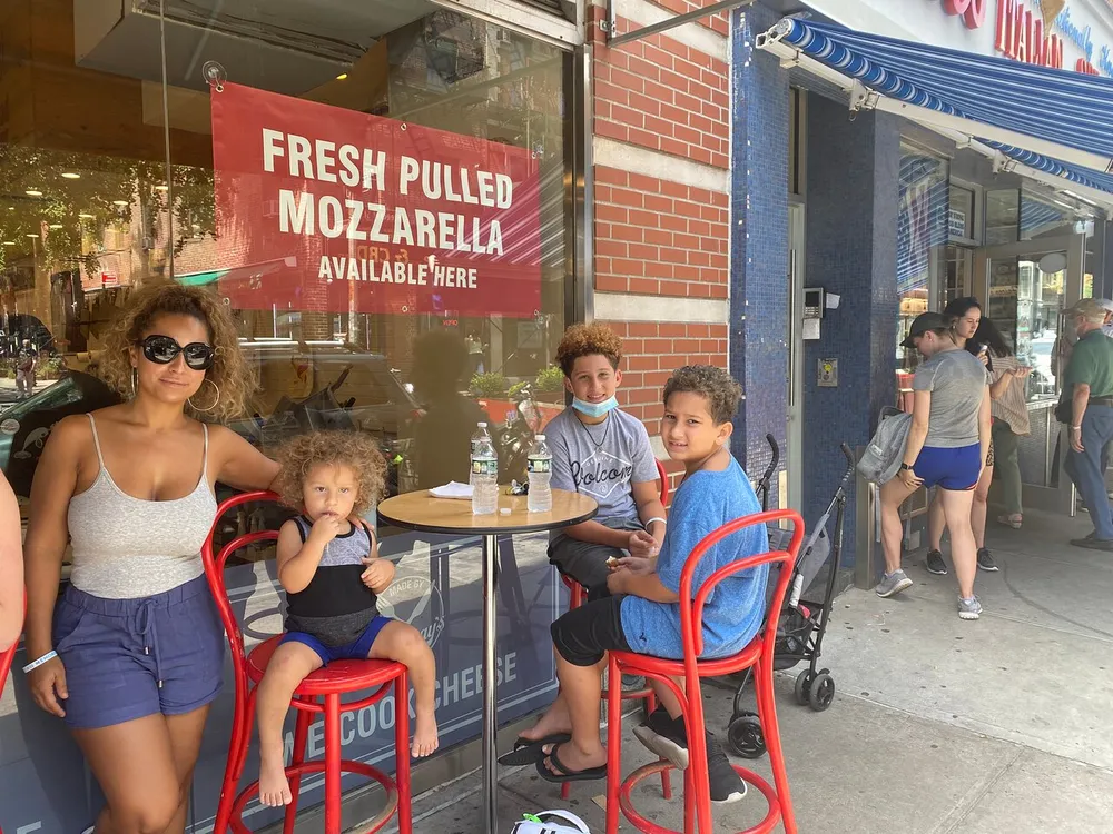 A group of people possibly a family is sitting at an outdoor dining table in front of a store advertising fresh pulled mozzarella