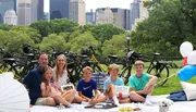A family of six is enjoying a picnic in a park with bicycles in the background, exuding a relaxed and happy atmosphere.