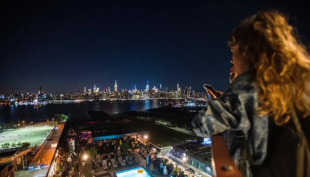 A person is holding a smartphone while enjoying a nighttime view of a vibrant city skyline from a high vantage point