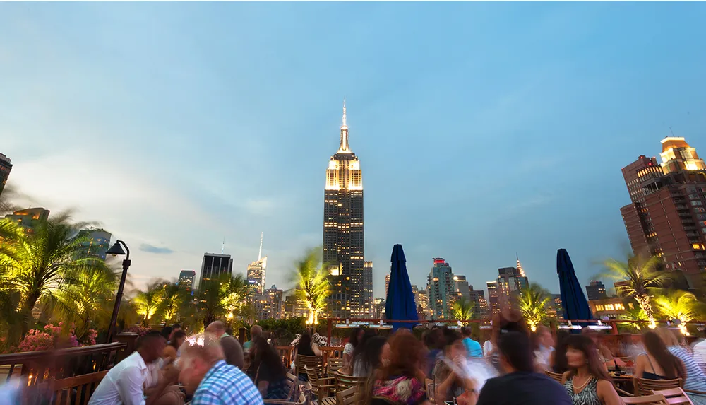 The image captures a bustling rooftop bar with people enjoying an evening against the backdrop of a lit-up Empire State Building and the New York City skyline at dusk