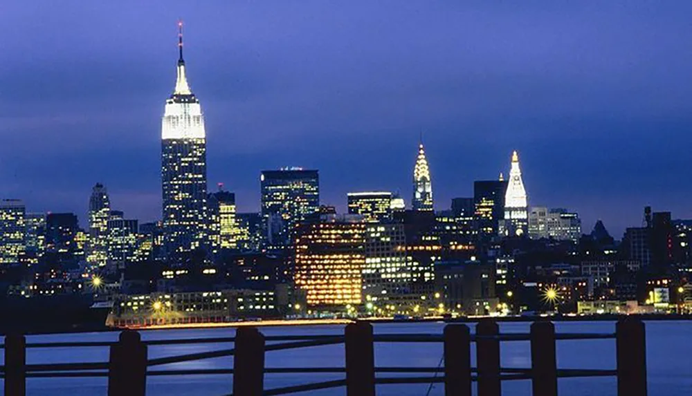 The image shows a twilight view of New York Citys iconic skyline with the Empire State Building prominently lit as seen from a waterside vantage point with a wooden railing in the foreground