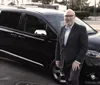 A smiling man in business attire is standing next to a black minivan in a parking area